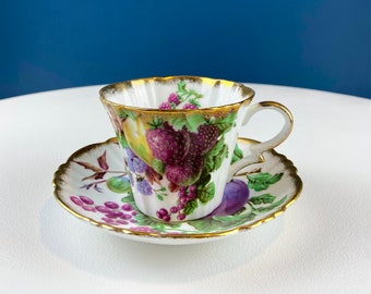 May include: A white teacup and saucer with a gold rim and a floral pattern of purple, green, and yellow fruits. The cup has a scalloped edge and a handle.
