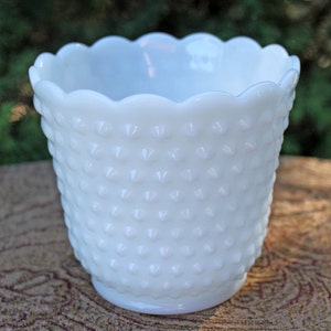May include: A white hobnail milk glass planter with a scalloped rim. The planter has a cylindrical shape and is covered in small, raised bumps. The interior is smooth and white. The planter is sitting on a patterned surface.
