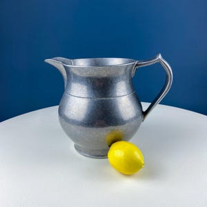 May include: A silver pitcher with a rounded body and a handle. The pitcher is sitting on a white surface next to a yellow lemon.