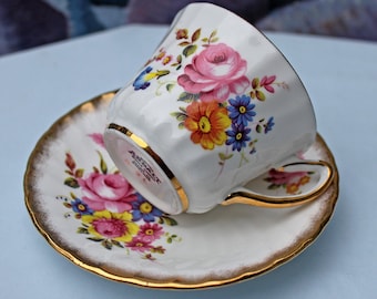 Puede incluir: Juego de taza y platillo blancos con un diseño floral que presenta rosas rosas, flores amarillas, naranjas y azules. El borde de la taza y el platillo están adornados con dorado. La taza de té está inclinada.