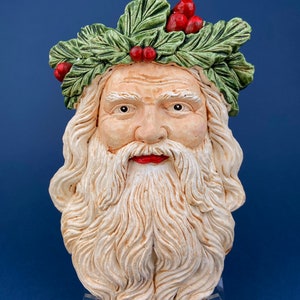 Puede incluir: Una cabeza tallada de Papá Noel con barba blanca y una corona de acebo verde con bayas rojas. La cara tiene rasgos detallados, incluyendo una boca roja y un tono de piel neutro. La escultura se exhibe sobre un soporte transparente.
