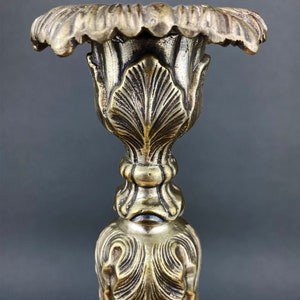 May include: A gold metal candlestick with an ornate design. The top of the candlestick is shaped like a flower with a wide, open mouth.