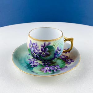 Könnte beinhalten: Vintage Tee-Set mit Tasse und Untertasse. Die Tasse und Untertasse sind mit handgemalten violetten Veilchen und grünen Blättern verziert. Die Ränder und der Henkel sind goldfarben. Der Hintergrund ist weiß und blau.