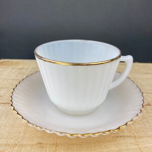 May include: A white milk glass teacup and saucer set with a gold trim. The cup has vertical ridges and the saucer has a scalloped edge.
