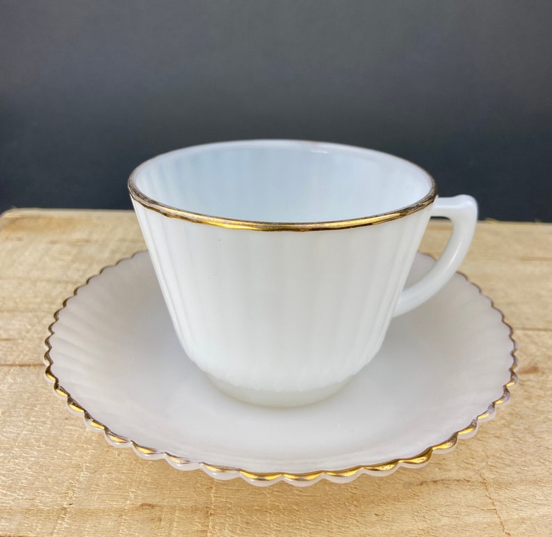 May include: A white milk glass teacup and saucer set with a gold trim. The cup has vertical ridges and the saucer has a scalloped edge.