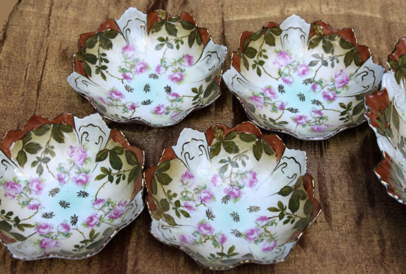 May include: Set of five white porcelain bowls with a floral design. The bowls have a scalloped edge and are decorated with pink roses, green leaves, and brown accents.