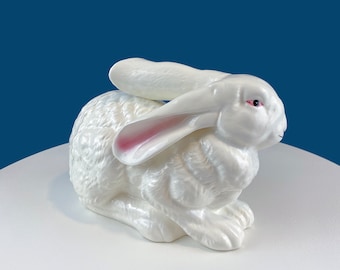 May include: A white ceramic rabbit figurine with pink ears. The rabbit is sitting on a white surface.