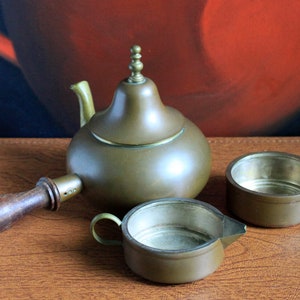 May include: A set of three antique brass tea service pieces, including a teapot with a wooden handle, a creamer, and a sugar bowl. The teapot has a decorative finial on the lid.