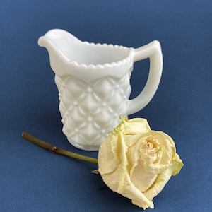 May include: A white milk glass creamer with a diamond pattern and a handle. A dried, cream-colored rose rests beside the creamer on a dark blue surface. The creamer has a scalloped rim.
