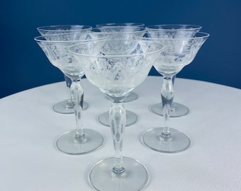 Op de afbeelding: Zes heldere, geëtste champagnecoupeglazen met bloemmotieven. De glazen hebben een brede, ondiepe kom, een slanke steel en een ronde voet. Ze staan op een wit oppervlak tegen een blauwe achtergrond.