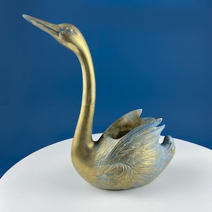 Puede incluir: Una escultura de cisne de latón con un cuello largo y curvado y plumas de ala detalladas. La escultura tiene un acabado dorado con toques de pátina azul-verdosa, y es probablemente un artículo decorativo.