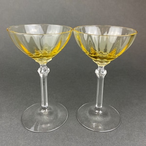 May include: Two clear glass champagne coupes with a yellow rim. The glasses are on a gray surface.