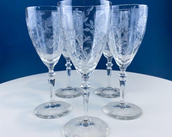 May include: Five clear wine glasses with etched floral designs. The glasses have a classic shape with a rounded bowl, a slender stem, and a circular base. The etched design features leaves and flowers. The glasses are arranged on a white surface against a blue background.