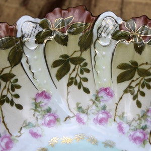 May include: A white porcelain bowl with a scalloped edge and a floral design. The bowl is decorated with green leaves, pink roses, and gold accents.