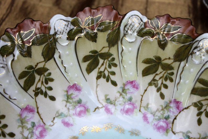May include: A white porcelain bowl with a scalloped edge and a floral design. The bowl is decorated with green leaves, pink roses, and gold accents.