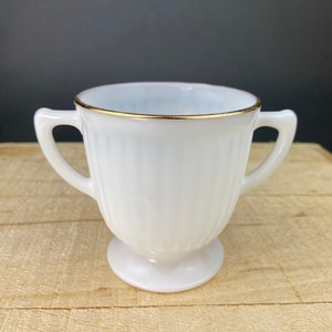 May include: White milk glass creamer with a gold rim and two handles. The creamer has a fluted design and a pedestal base.