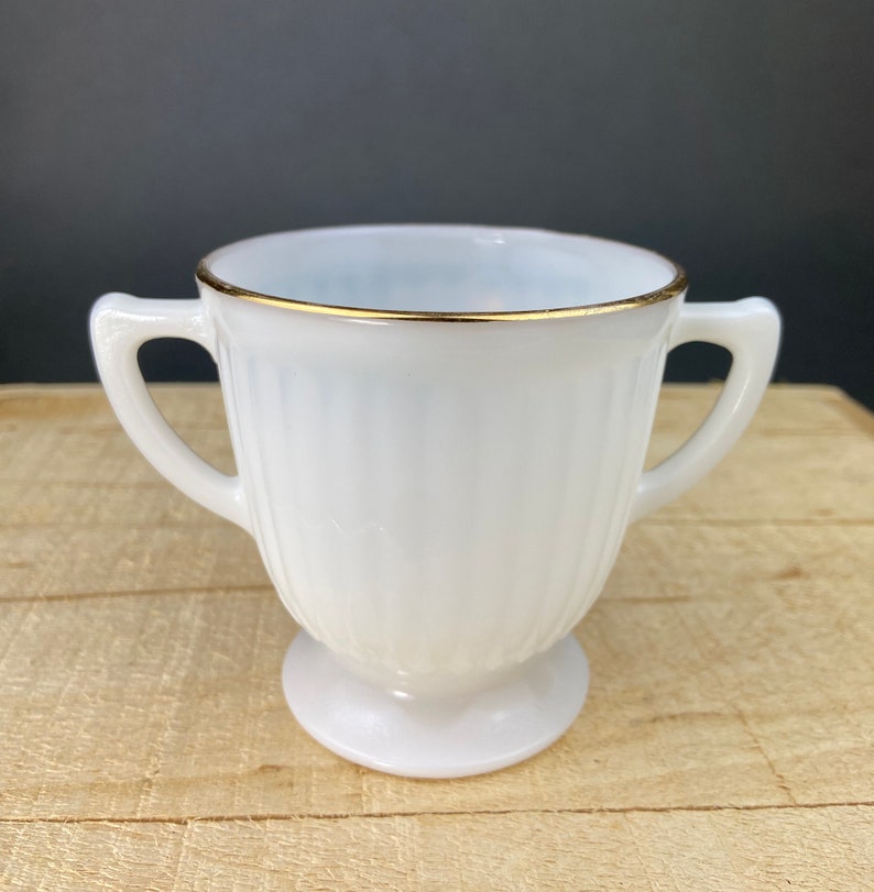 May include: White milk glass creamer with a gold rim and two handles. The creamer has a fluted design and a pedestal base.