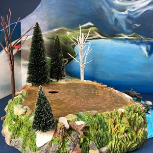 May include: A miniature winter scene with a snowy, rocky base, a small evergreen tree, and a brown dirt path. The scene is set against a backdrop of a blue lake and a snow-capped mountain.