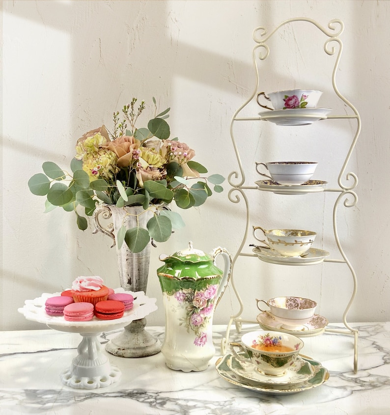 May include: A white three-tiered serving stand with a floral design, holding vintage teacups and saucers. A bouquet of pink and white roses sits in a white vase on a marble surface. A white cake stand with pink and red macarons sits in front of the stand.