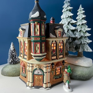 May include: A miniature ceramic coffee shop building with a tall dark tower, brick-textured walls, and illuminated windows. The structure features a sign reading Coffee Shop above the front door, with a small figurine of a person holding a tree.