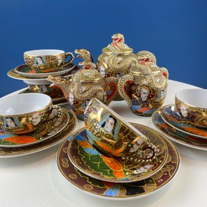 Puede incluir: Un juego de té dorado con diseños de dragones y retratos, que incluye una tetera, azucarero y tazas con platillos. El juego está dispuesto sobre una superficie blanca sobre un fondo azul. Las tazas y platillos están decorados con retratos.