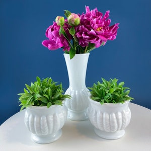 Puede incluir: Tres jarrones de cerámica blanca con una superficie texturizada. El jarrón del centro es más alto y contiene un ramo de peonías rosas. Los dos jarrones más pequeños a cada lado contienen plantas verdes.