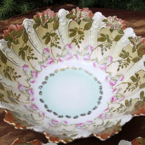 May include: A large, white porcelain bowl with a scalloped edge. The bowl is decorated with a floral pattern of pink roses and green leaves. The bowl has a light blue center and a gold rim.