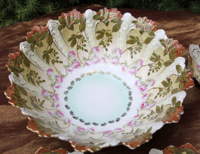 May include: A large, white porcelain bowl with a scalloped edge. The bowl is decorated with a floral pattern of pink roses and green leaves. The bowl has a light blue center and a gold rim.