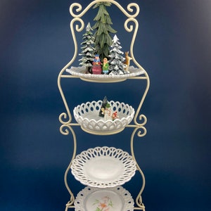 May include: A white metal tiered stand with three white ceramic plates. The top plate has a small Christmas scene with trees, a snowman, and a deer. The stand is perfect for displaying holiday treats or decorations.