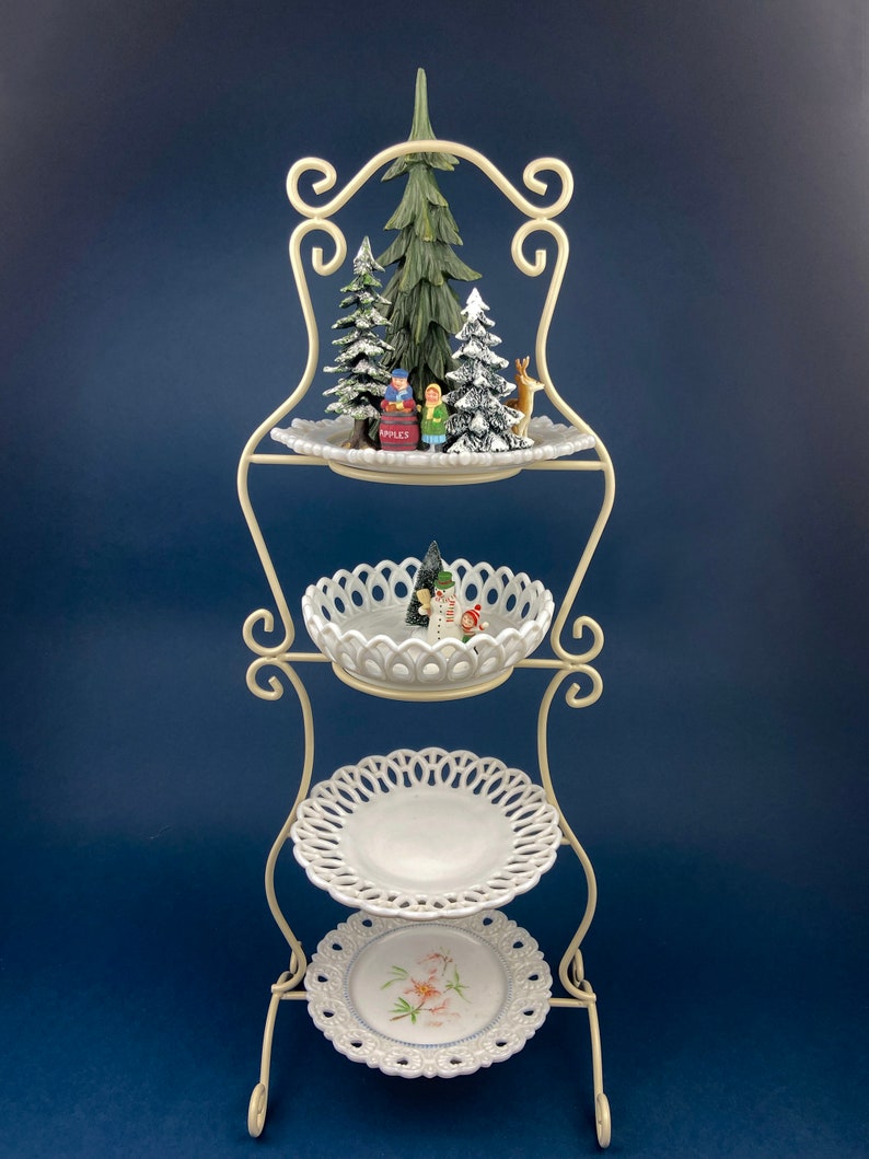 May include: A white metal tiered stand with three white ceramic plates. The top plate has a small Christmas scene with trees, a snowman, and a deer. The stand is perfect for displaying holiday treats or decorations.