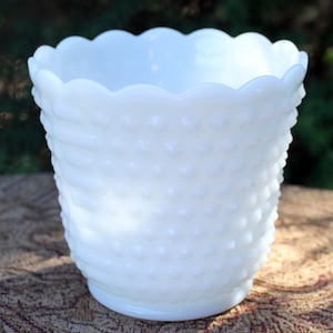 May include: A white hobnail milk glass planter with a scalloped rim. The cylindrical vase features a textured surface with raised dots and vertical ribbing. The vintage-style container is suitable for indoor or outdoor use.