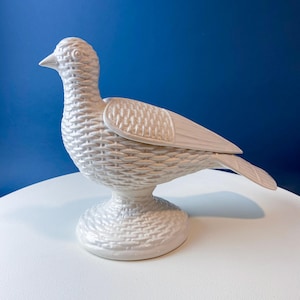 May include: A white ceramic bird figurine with a textured, woven basket-like pattern. The bird is perched on a round base and has detailed wings and tail feathers. The background is a solid blue.