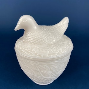 Op de afbeelding: Witte melkglas hen en nestvormige snoepschotel. De hen zit op een nest van bladeren en heeft een gedetailleerd, getextureerd oppervlak.