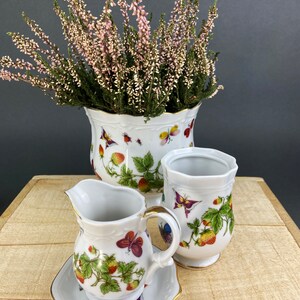 May include: A white porcelain creamer and sugar set with a floral and butterfly design. The set features a creamer with a handle, a sugar bowl, and a matching saucer. The design includes strawberries, leaves, and butterflies in shades of pink, purple, and blue.