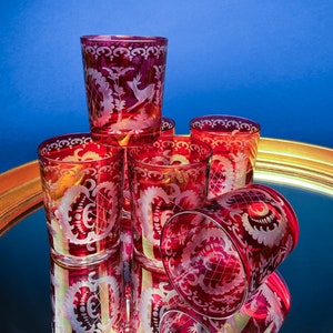 Puede incluir: Juego de seis vasos rojos de vidrio con dise&ntilde;os florales y animales grabados. Los vasos est&aacute;n dispuestos en c&iacute;rculo sobre una bandeja dorada con superficie de espejo.