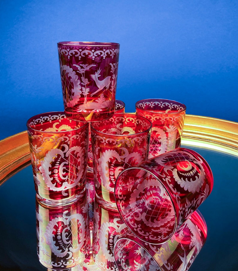 Puede incluir: Juego de seis vasos rojos de vidrio con dise&ntilde;os florales y animales grabados. Los vasos est&aacute;n dispuestos en c&iacute;rculo sobre una bandeja dorada con superficie de espejo.