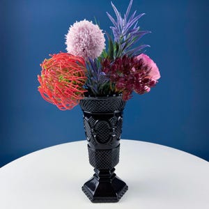 Könnte beinhalten: Eine schwarze, strukturierte Glasvase mit quadratischem Sockel hält einen Strauß aus rosa, roten und violetten Blumen. Die Vase steht auf einer weißen Oberfläche vor einem blauen Hintergrund.