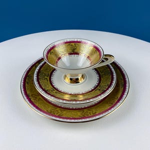 Puede incluir: Un juego de taza y platillo de té blanco con un patrón floral dorado y burdeos. La taza y el platillo tienen un borde dorado y un delicado diseño floral.