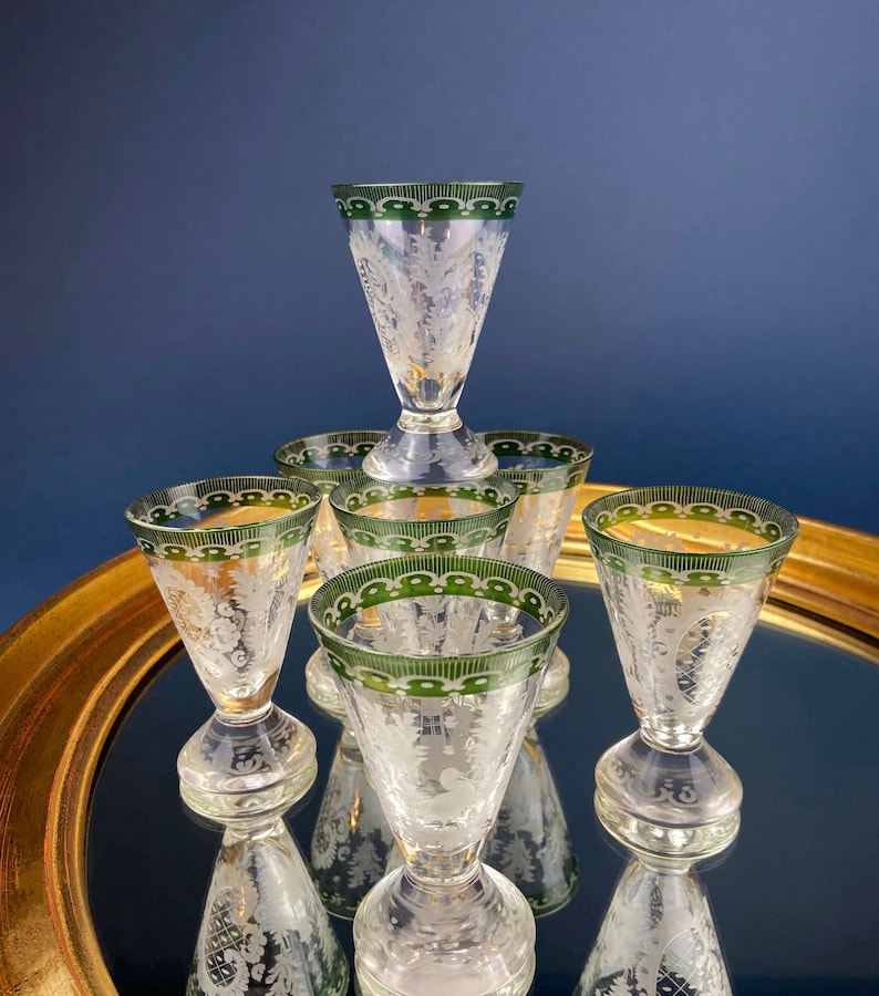 Puede incluir: Conjunto de seis vasos de chupito de vidrio transparente con borde verde y dise&ntilde;o floral grabado. Los vasos est&aacute;n dispuestos sobre una superficie de espejo dorado.