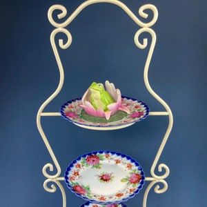 May include: White metal tiered stand with three porcelain plates. The top plate features a green frog figurine sitting in a pink flower. The bottom two plates have a blue and white floral design.