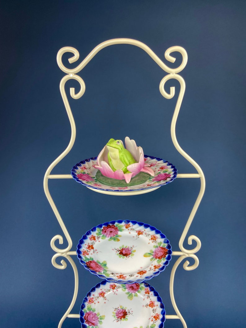 May include: White metal tiered stand with three porcelain plates. The top plate features a green frog figurine sitting in a pink flower. The bottom two plates have a blue and white floral design.