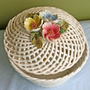 Puede incluir: Cesta de cerámica blanca con tapa tejida decorada con tres flores de colores. Las flores son amarillas, azules y rosas.