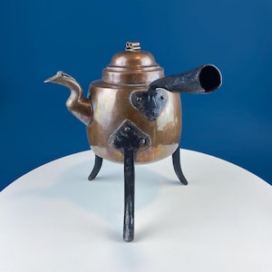 May include: A copper teapot with a black iron spout and legs. The teapot has a domed lid and a decorative handle.