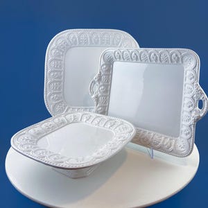 Puede incluir: Tres fuentes blancas de cerámica para servir con bordes ornamentados con motivos florales y volutas en relieve. Las fuentes son cuadradas y rectangulares, una de ellas con asas. Están dispuestas sobre una superficie blanca sobre un fondo azul.