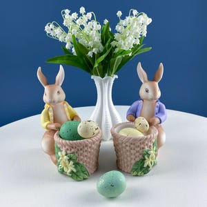 May include: Two ceramic Easter bunny figurines, each holding a basket of speckled eggs. The bunnies wear jackets, one yellow and one purple. A white vase with white flowers sits between them. A single speckled egg rests in front.