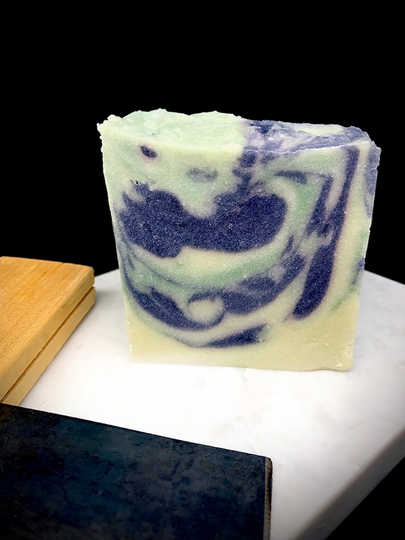 Puede incluir: Una pastilla de jab&oacute;n artesanal con un patr&oacute;n de remolino en tonos de verde, blanco y morado. El jab&oacute;n est&aacute; sobre una superficie de m&aacute;rmol blanco.
