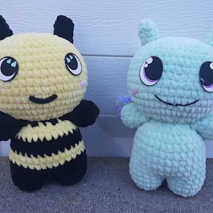 Puede incluir: Dos juguetes de peluche tejidos a crochet. Uno es una abeja amarilla y negra con una cara sonriente y ojos grandes. El otro es un juguete de peluche azul claro con una cara sonriente y ojos grandes. Ambos juguetes tienen una flor tejida a crochet en el brazo.