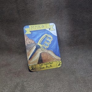 May include: A rectangular card with a hand-drawn illustration of an ice pick. The card features the text "MMK&T" above a blue and brown landscape with a gold ice pick. The text "Les Apaches Ice Pick" is also present.