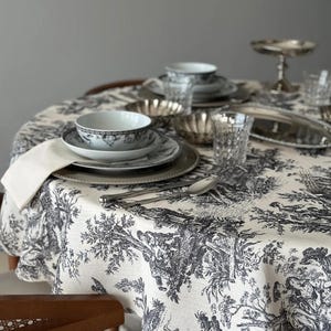 Könnte beinhalten: Ein gedeckter Esstisch. Der Tisch ist mit einer weißen Tischdecke mit schwarzem Toile-de-Jouy-Muster bedeckt. Silberbesteck, Teller, Schalen und Gläser sind auf dem Tisch angeordnet. Eine weiße Serviette liegt auf dem Tisch.