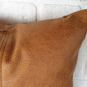 May include: A brown faux leather pillow with a textured surface. The pillow is shown against a white brick wall.